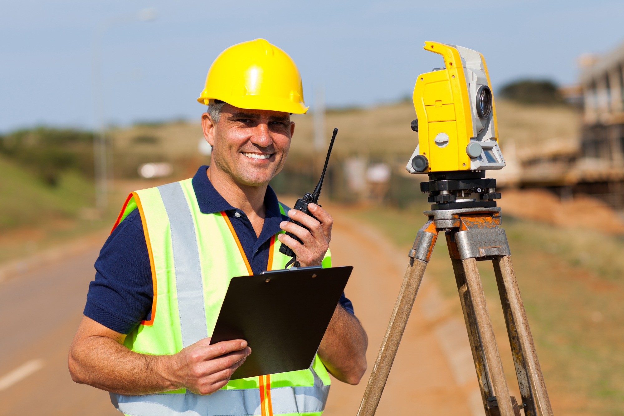 Surveyor in a yellow hard hat holds a clipboard and radio, standing next to a surveying instrument on a construction site.